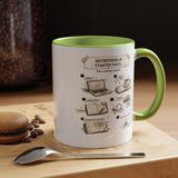 A white ceramic coffee mug with a green handle and interior, featuring a printed 'Entrepreneur Starter Pack' assembly-style graphic on the front. The mug sits on a wooden surface with a spoon in front and coffee beans nearby, against a dark background.